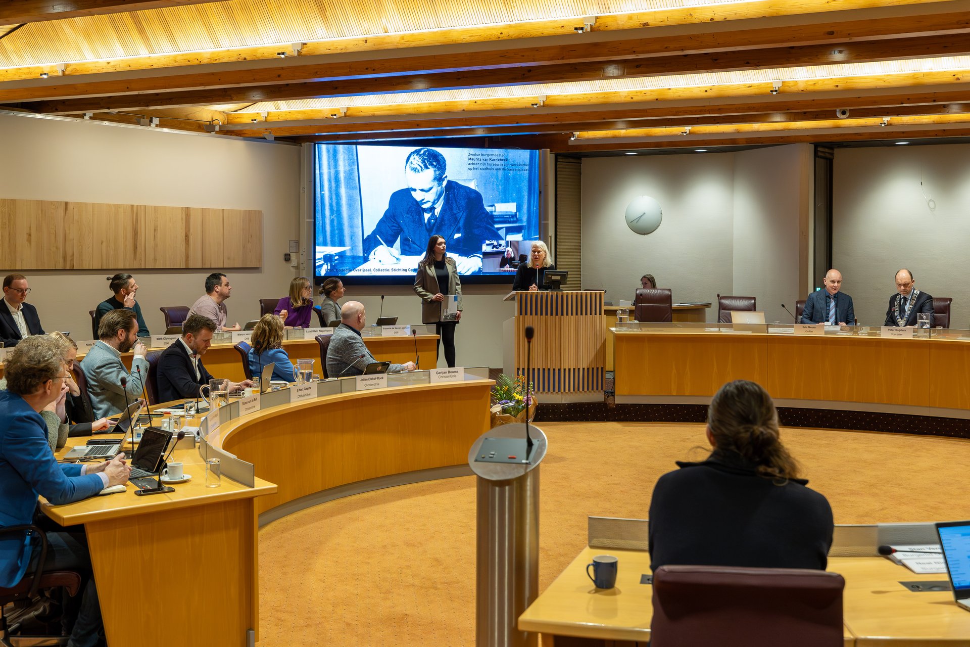 Een afbeelding van de raadzaal van het moment dat het NIOD het rapport overdraagt aan de burgemeester.