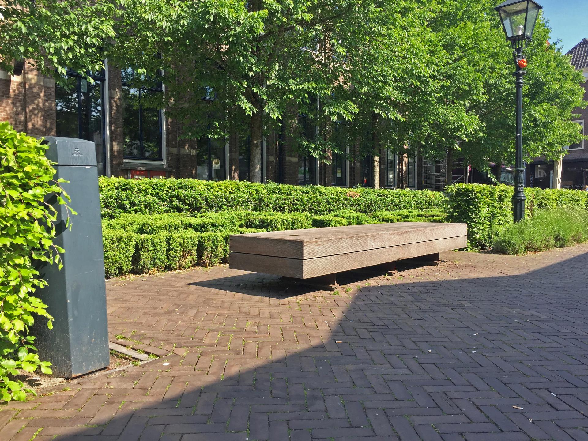 De foto toont een zitbank zonder leuningen op het plein de Nieuwe Markt in Zwolle. De bank is omgeven door heggen en verlichting..